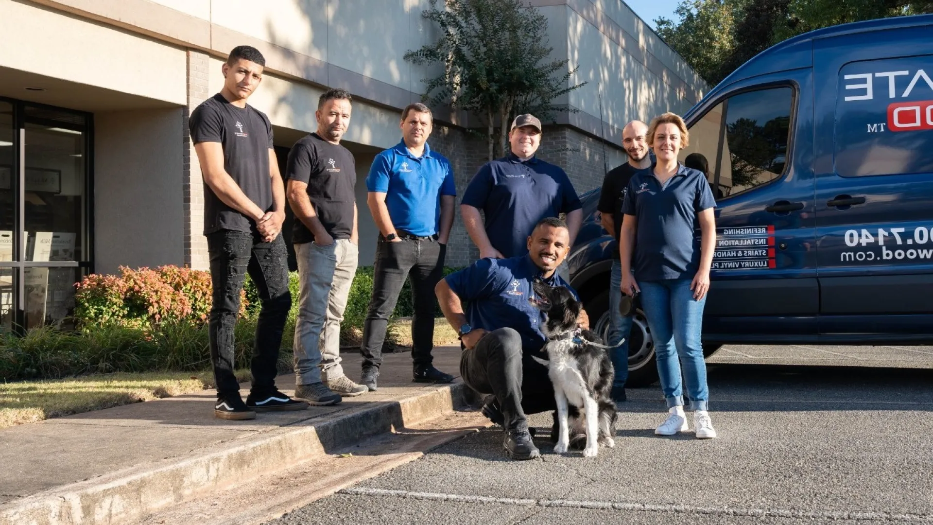 Team of seven workers with a dog posing outside a service van and building on a sunny day