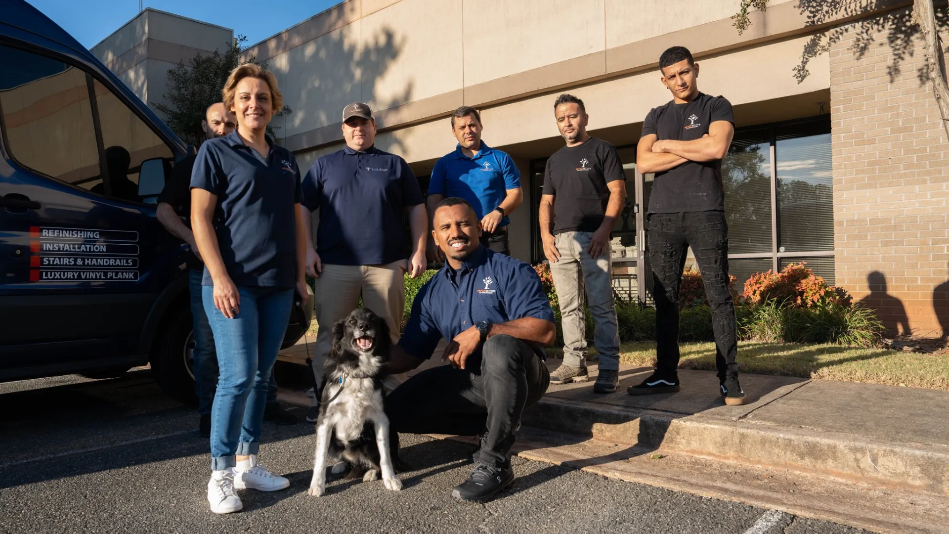 Group of six workers and a dog posing outside a building near a work van on a sunny day