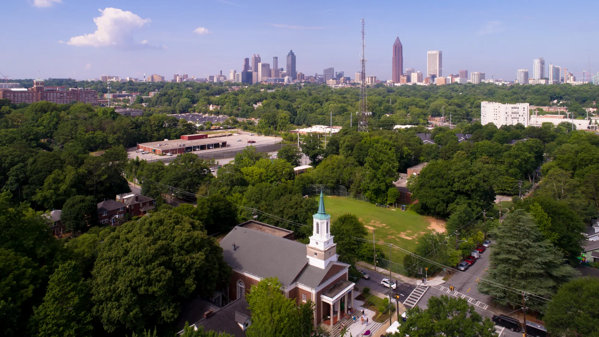 Aerial view of Atlanta skyline with dense green trees and a church with a steeple in the foreground on a sunny day.