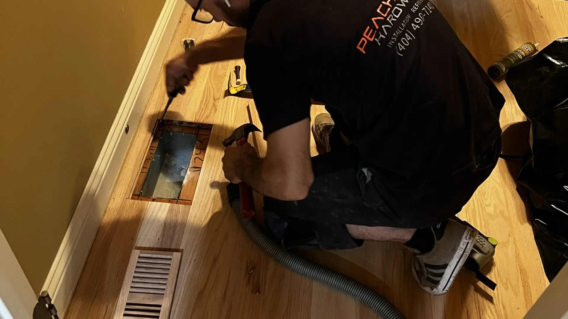 Worker installs or repairs hardwood flooring around a vent in a small room next to a pedestal sink.