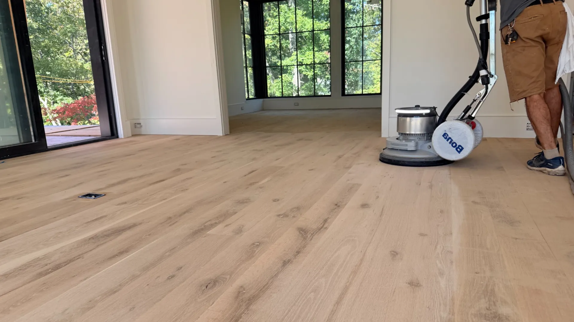 Person sanding and refinishing a wood floor in a bright room with large windows and natural light.