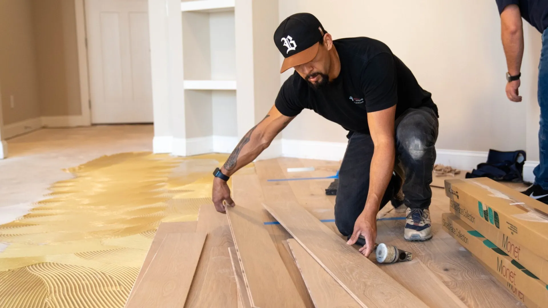 Two men installing wooden flooring in a room with adhesive spread on the floor.