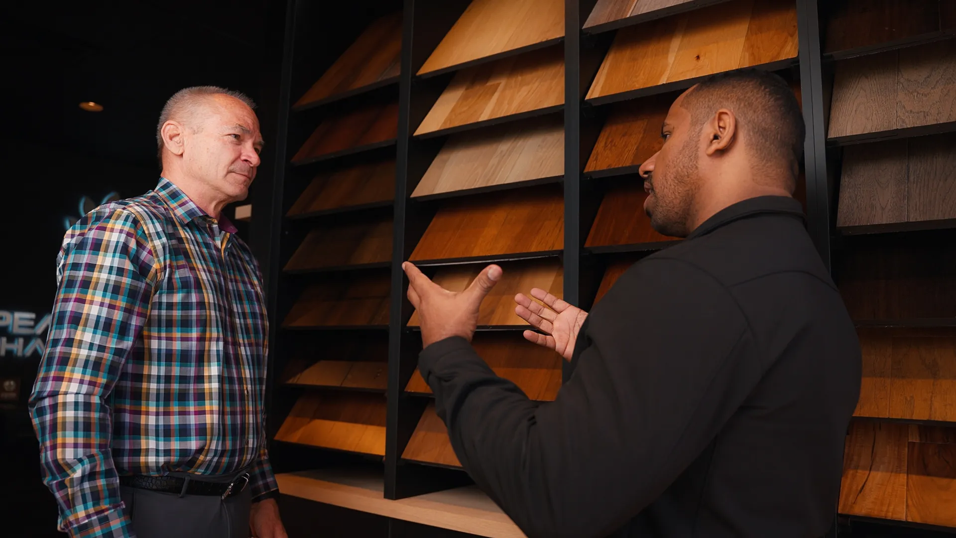 Two men discussing wooden floor samples displayed on a wall in a showroom or store.