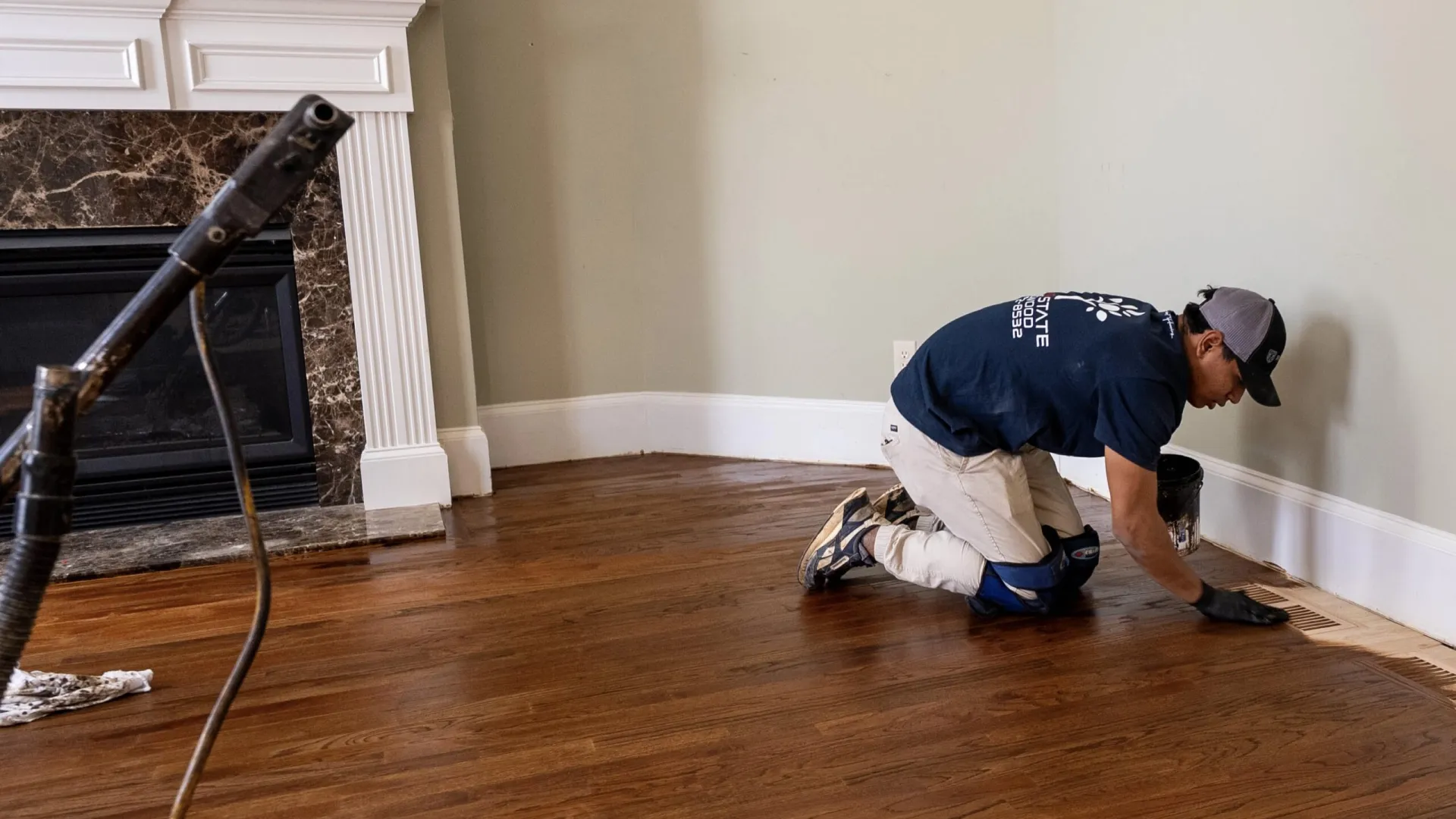 Professional worker refinishing hardwood floor near fireplace in a home interior, applying stain along the edges.