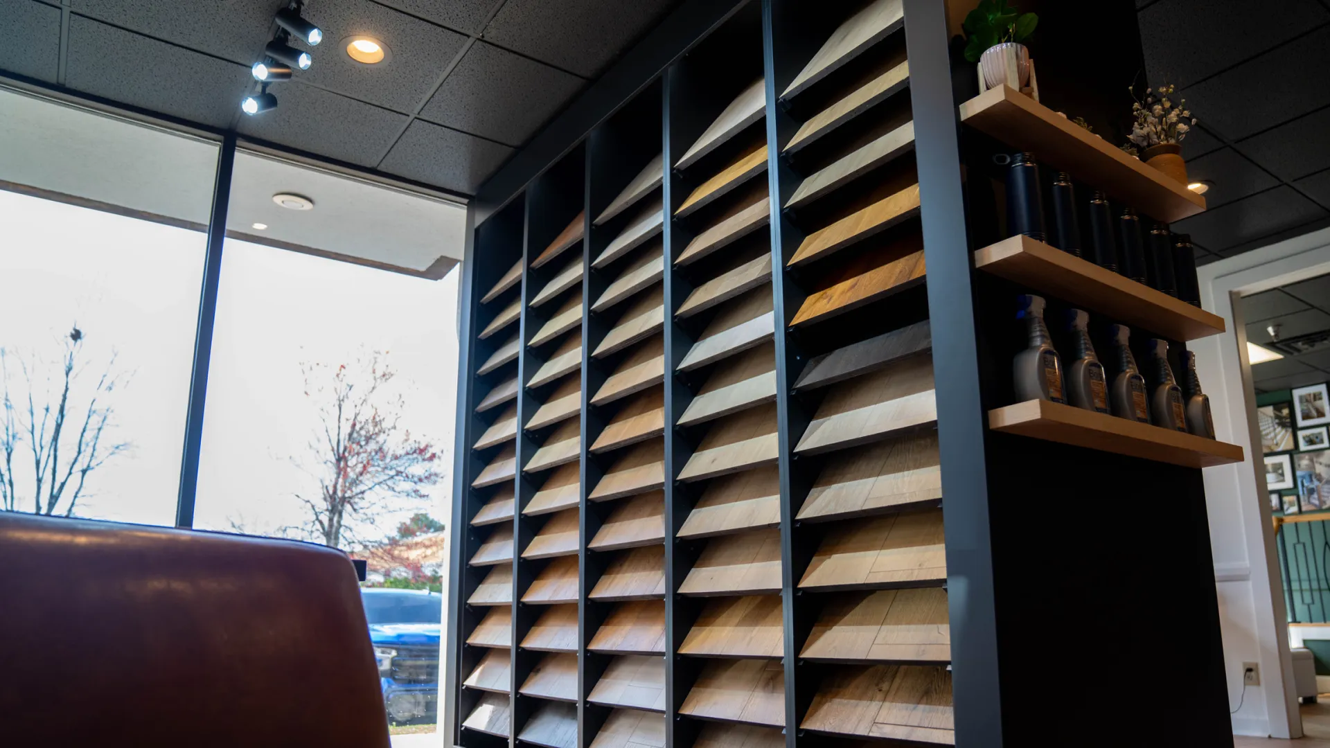 Modern interior with wood sample display, leather chair, large windows, and decorative shelves with bottles and plants.