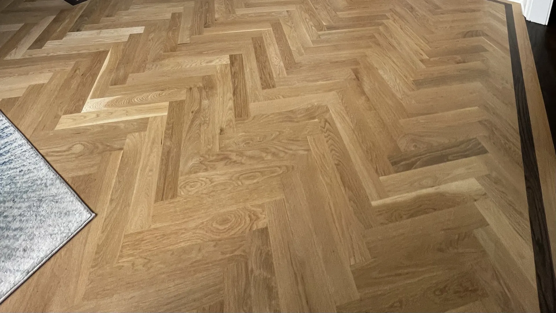 Light wood parquet flooring in a herringbone pattern with a dark wood border and a gray rug corner.