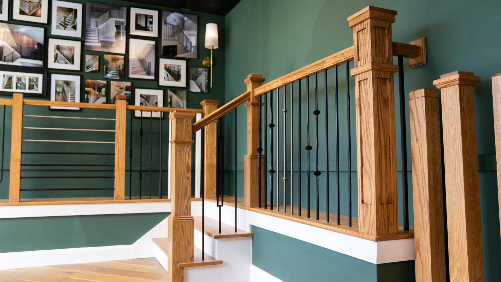 Modern wooden staircase with black metal balusters and green walls decorated with framed photographs and a sconce light