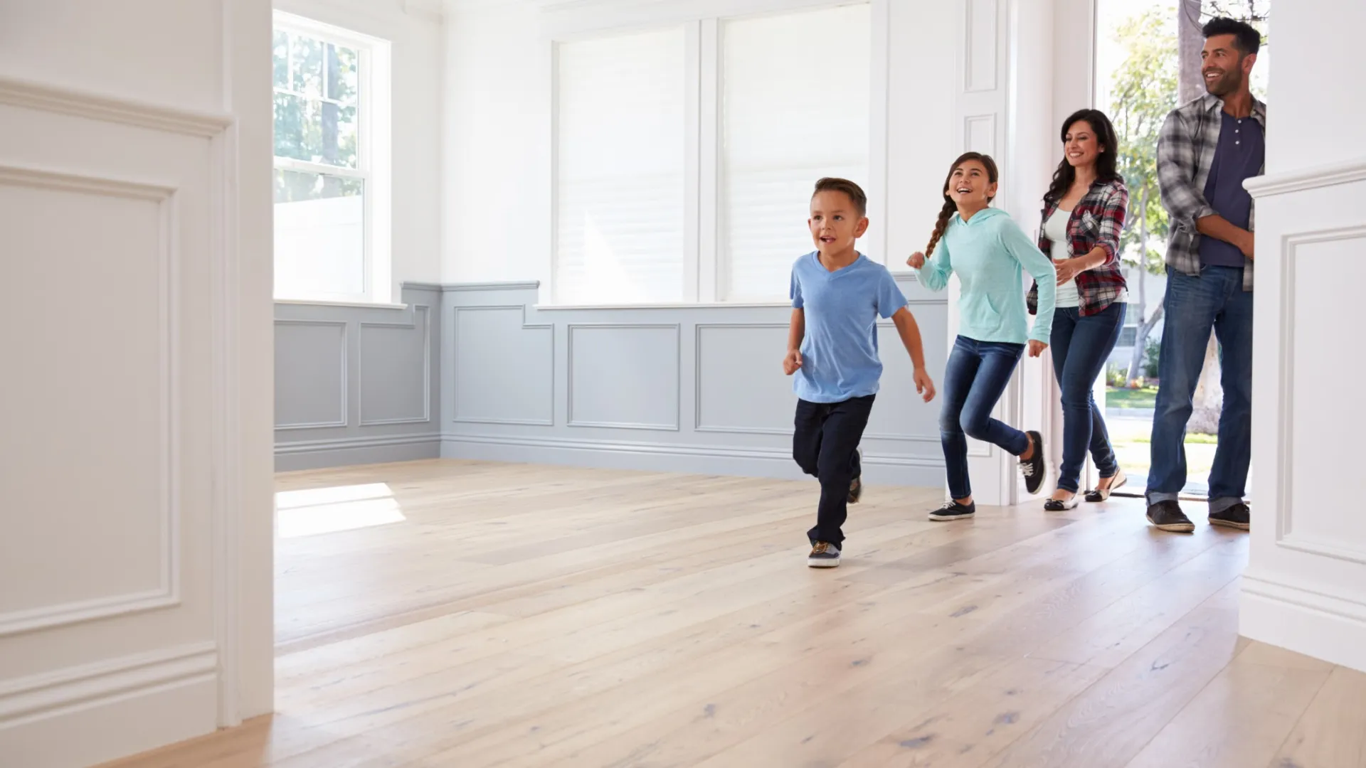 Happy family entering a bright empty room with wooden floors, excited children running ahead of parents