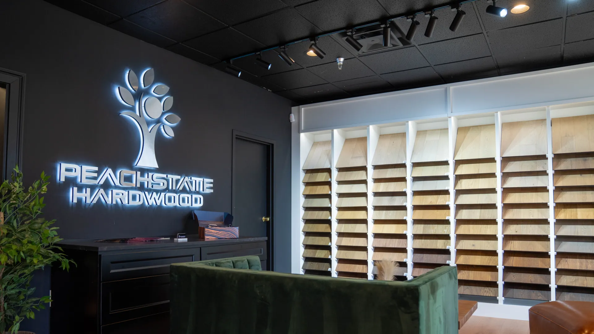 Modern showroom with illuminated Peachstate Hardwood logo, hardwood floor samples, and green velvet couch.