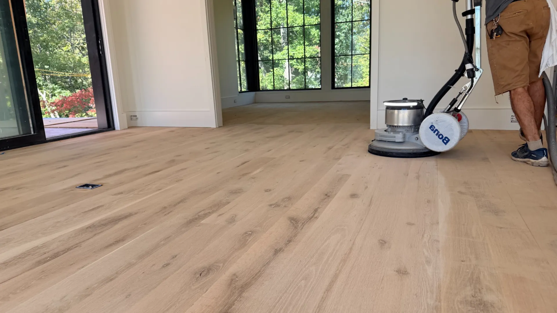 Worker refinishing light wooden floor with buffer machine in bright modern room with large windows.