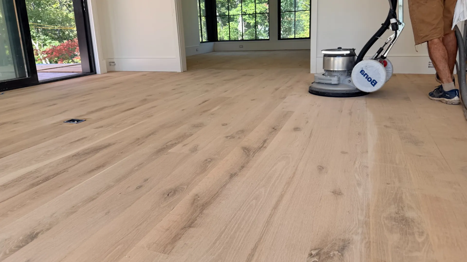Person sanding light wood flooring in a bright room with large windows and sliding glass door.