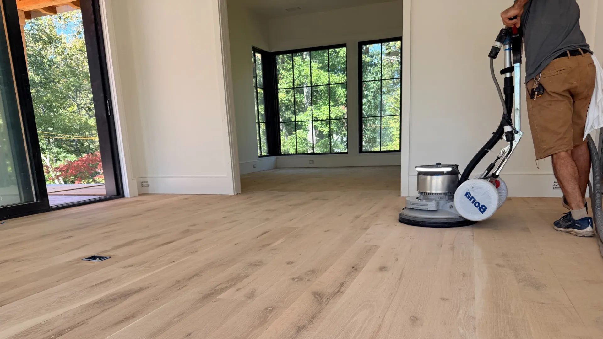 Worker refinishing light wooden floor with buffer machine in bright modern room with large windows.