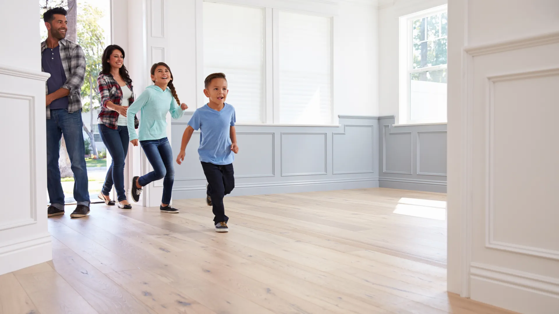 Happy family entering a bright empty room with wooden floors, excited children running ahead of parents