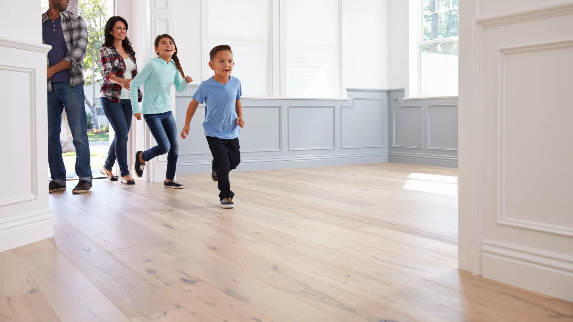 Happy family entering a bright empty room with wooden floors, excited children running ahead of parents