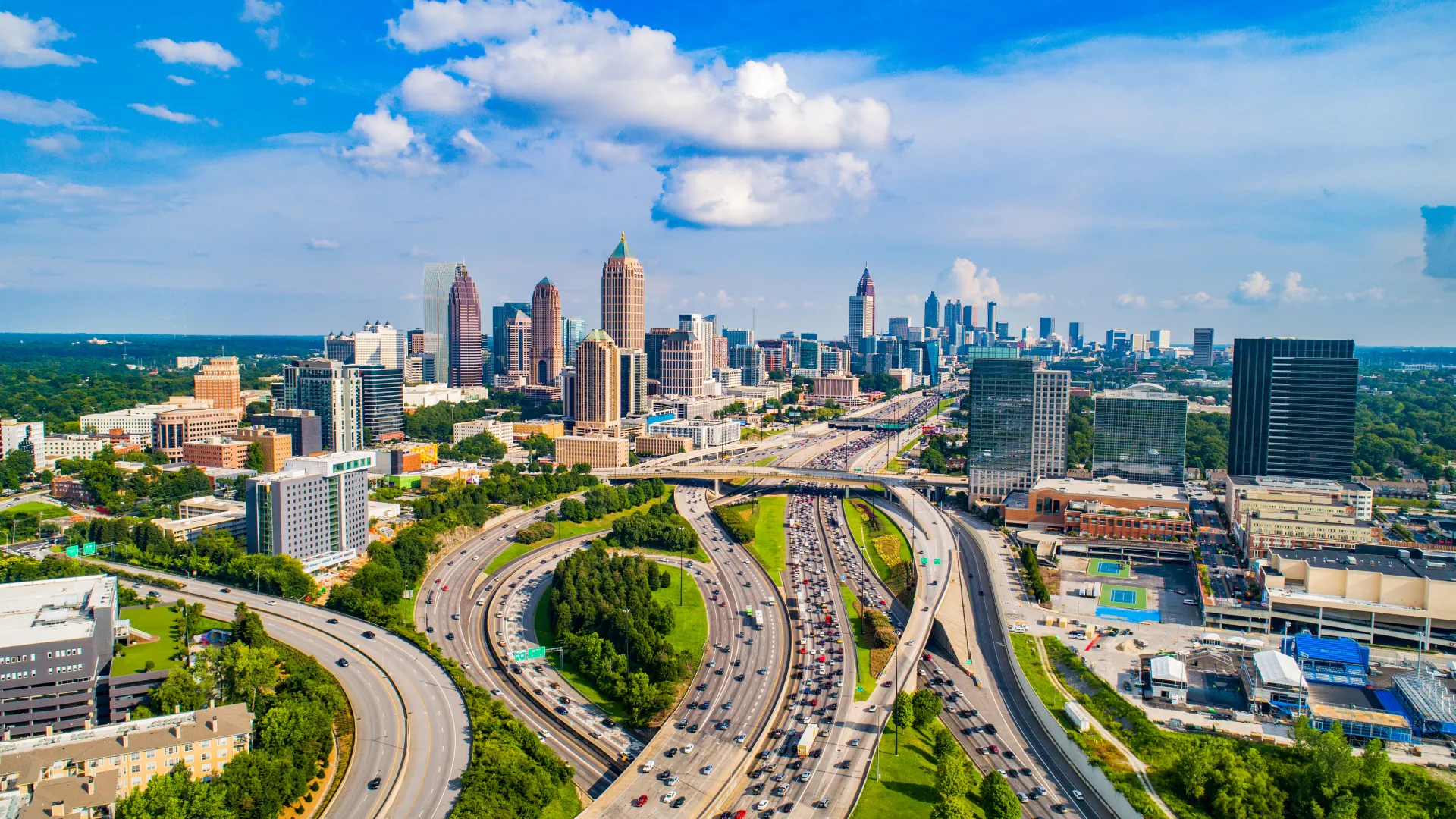Aerial view of a bustling city skyline with multiple highways merging and traffic under a blue sky with clouds.