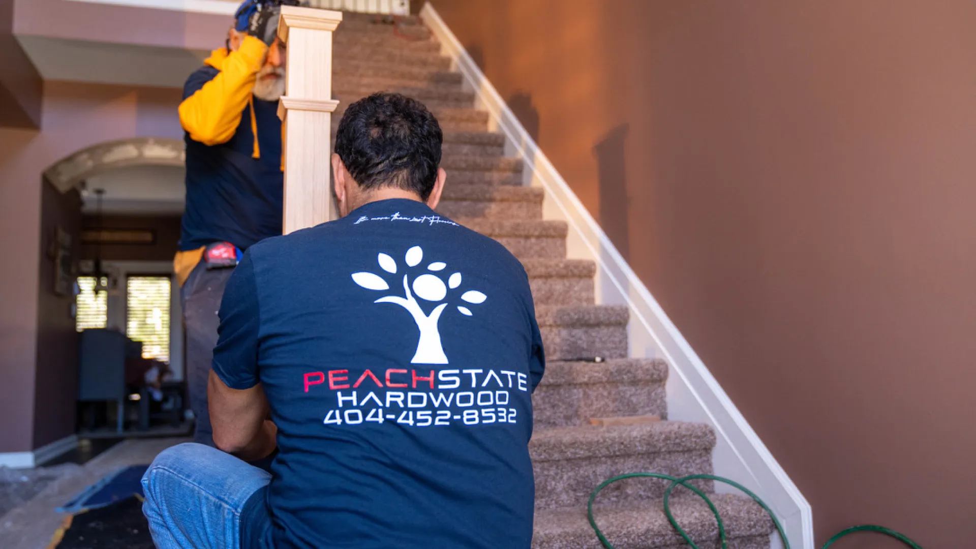 Two workers installing new hardwood stair railing in a home interior with Peach State Hardwood branding visible.