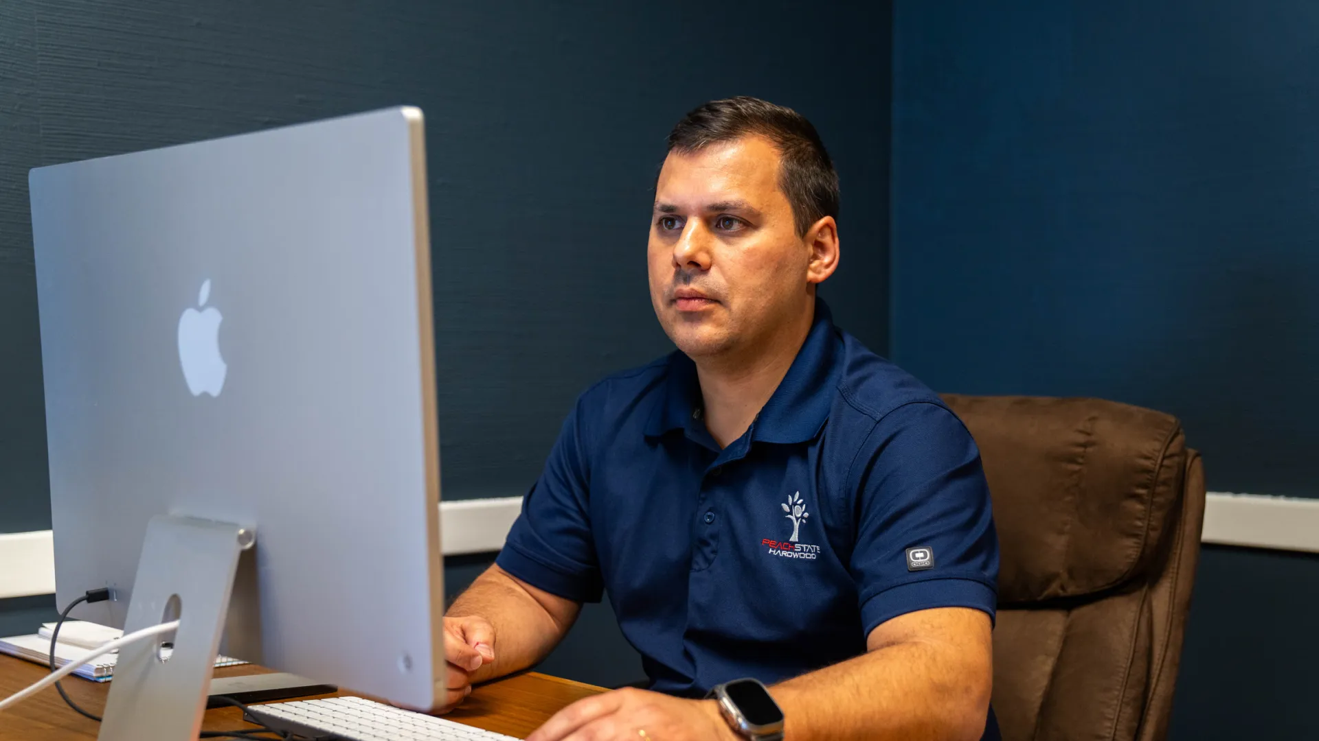 Man in navy blue polo working on an Apple iMac at a wooden desk in a dark office environment