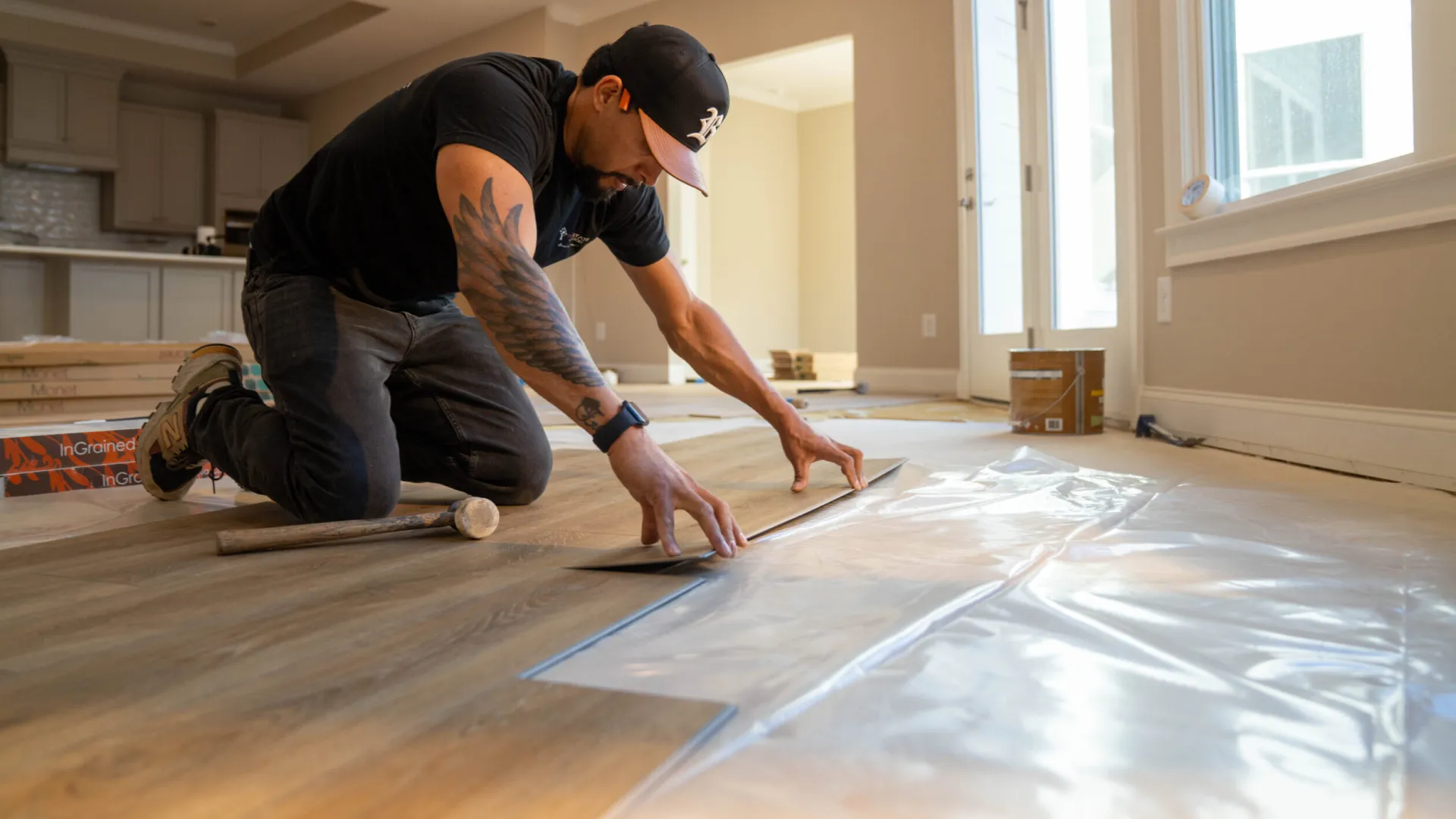 Man installing wooden flooring over plastic underlayment in a sunlit room with tools nearby.