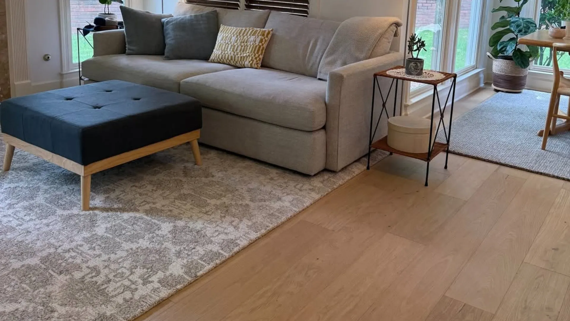 Modern living room with beige sofa, blue ottoman, wooden floor, patterned rug, and natural light from large windows.