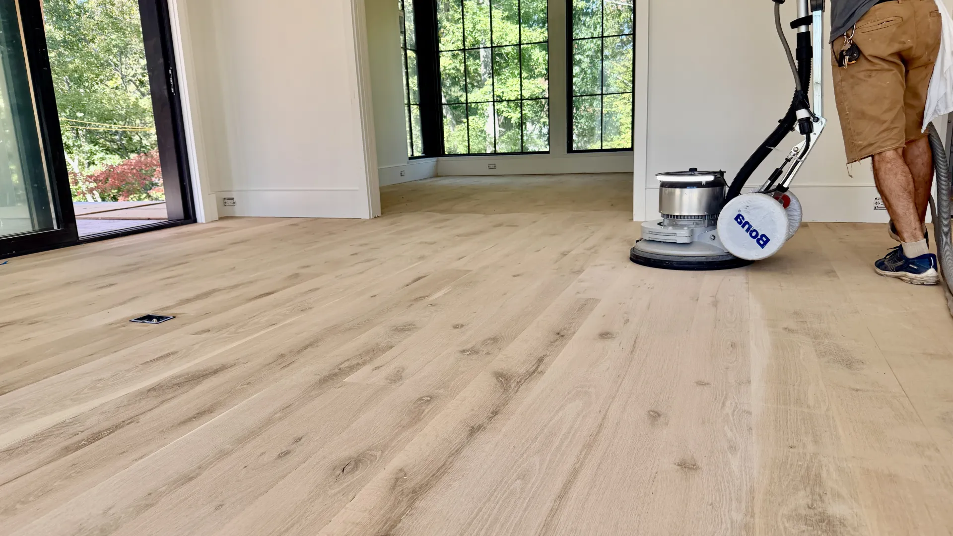 Person polishing light hardwood floor using a floor buffer in a bright room with large windows.