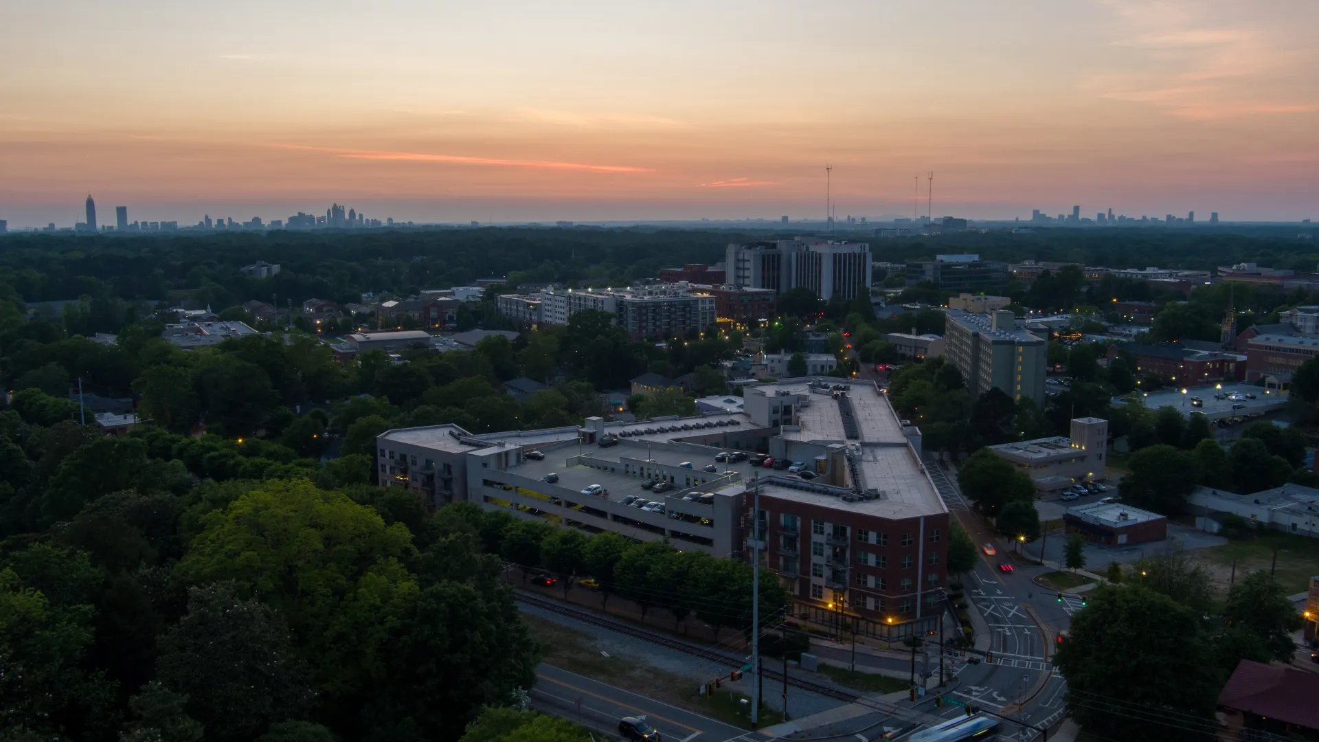 Aerial view of a cityscape at sunset with buildings, roads, and lush green trees under a colorful sky.