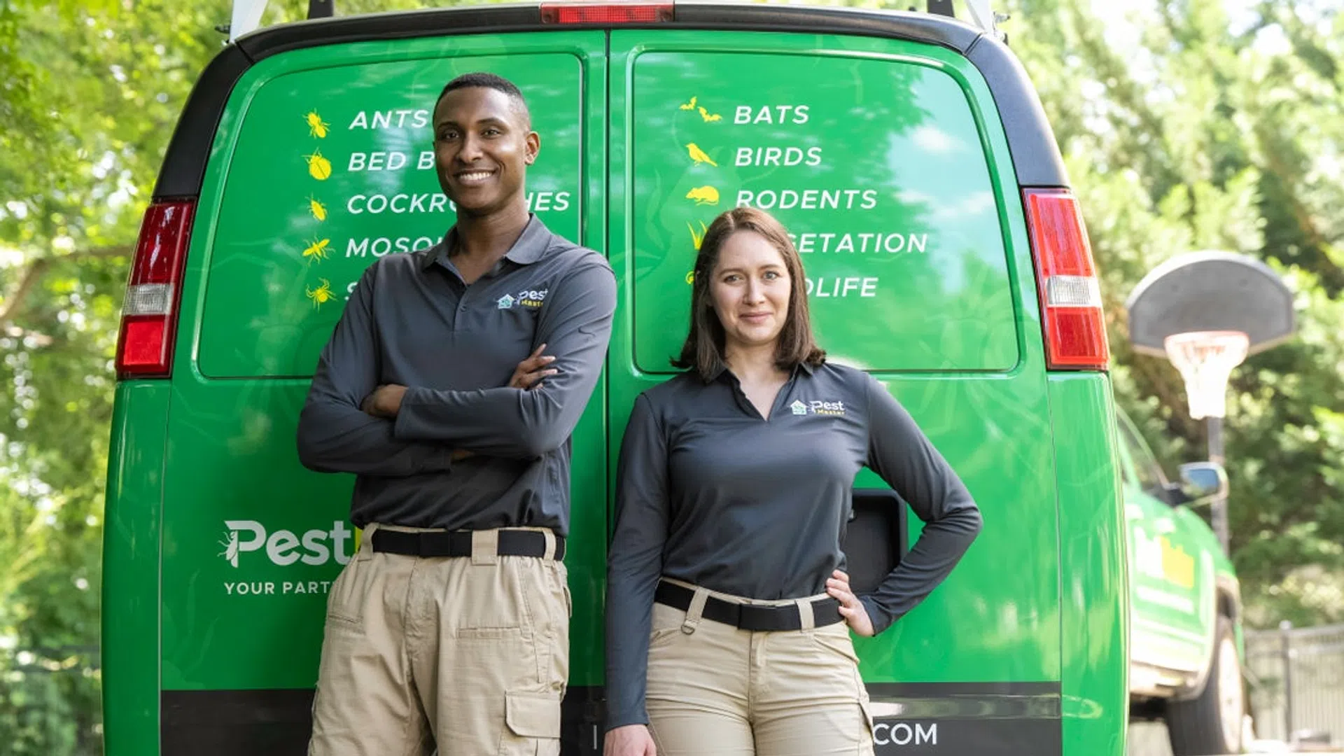 Two pest control workers in uniforms standing in front of green company van with pest service listings.