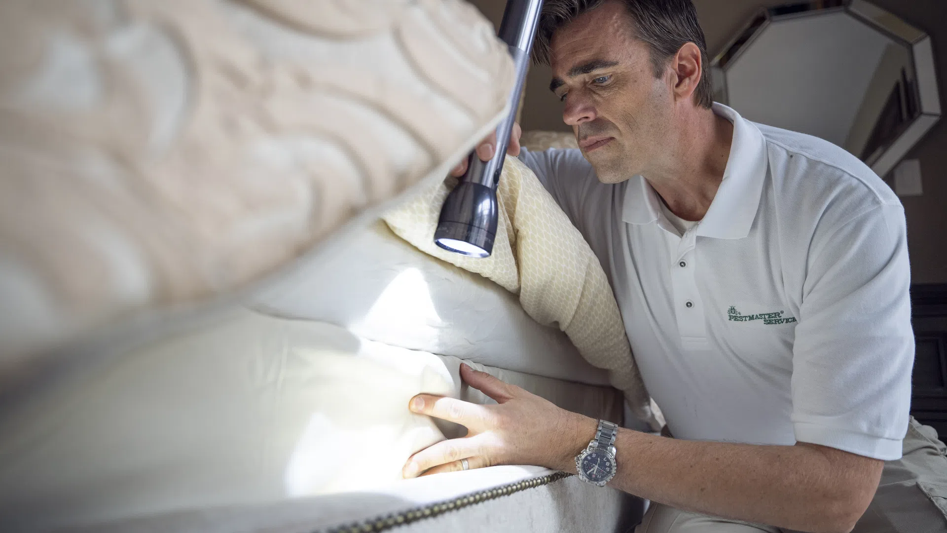 PestMaster Central Houston technician inspects mattress with flashlight for bed bugs or pests in a bedroom.