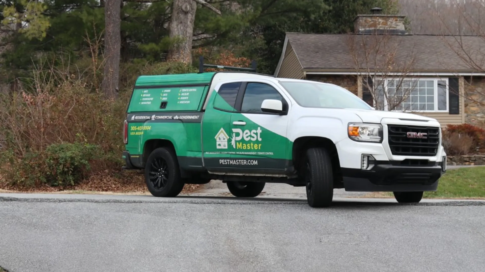 Green and white pest control truck parked on a driveway in front of suburban houses with trees and lawns.