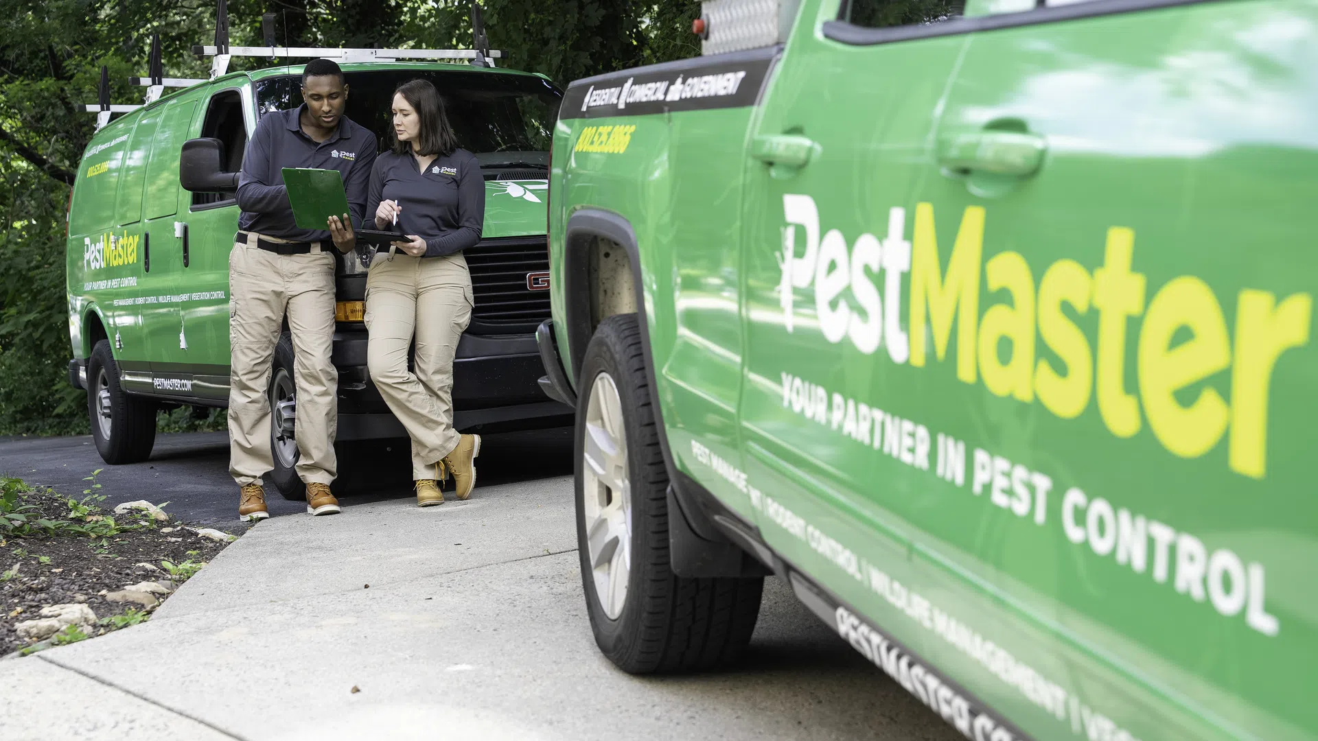 Two PestMaster pest control workers reviewing tablet near branded green service vehicles in residential driveway