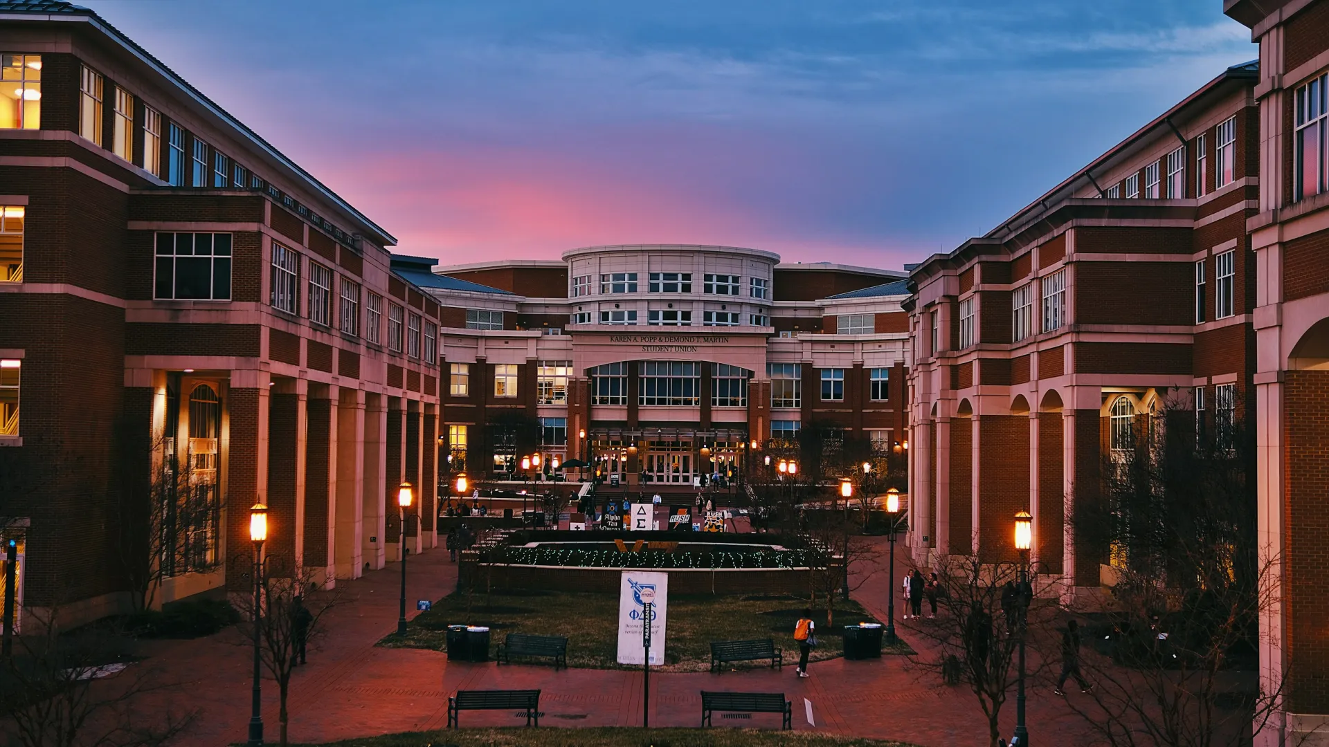 University campus courtyard at dusk with modern brick buildings and lit lampposts under a colorful sky