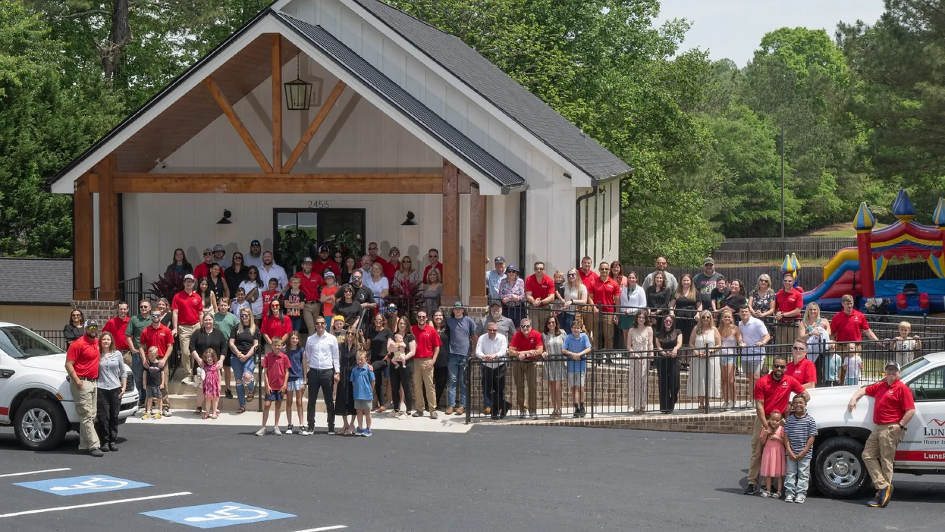 Large group of people posing in front of a modern building with two white company trucks in a parking lot surrounded by trees