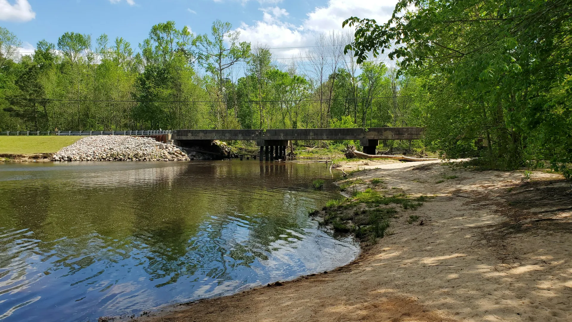 Sandy riverbank with clear water, trees, and a concrete bridge under blue sky with clouds on a sunny day