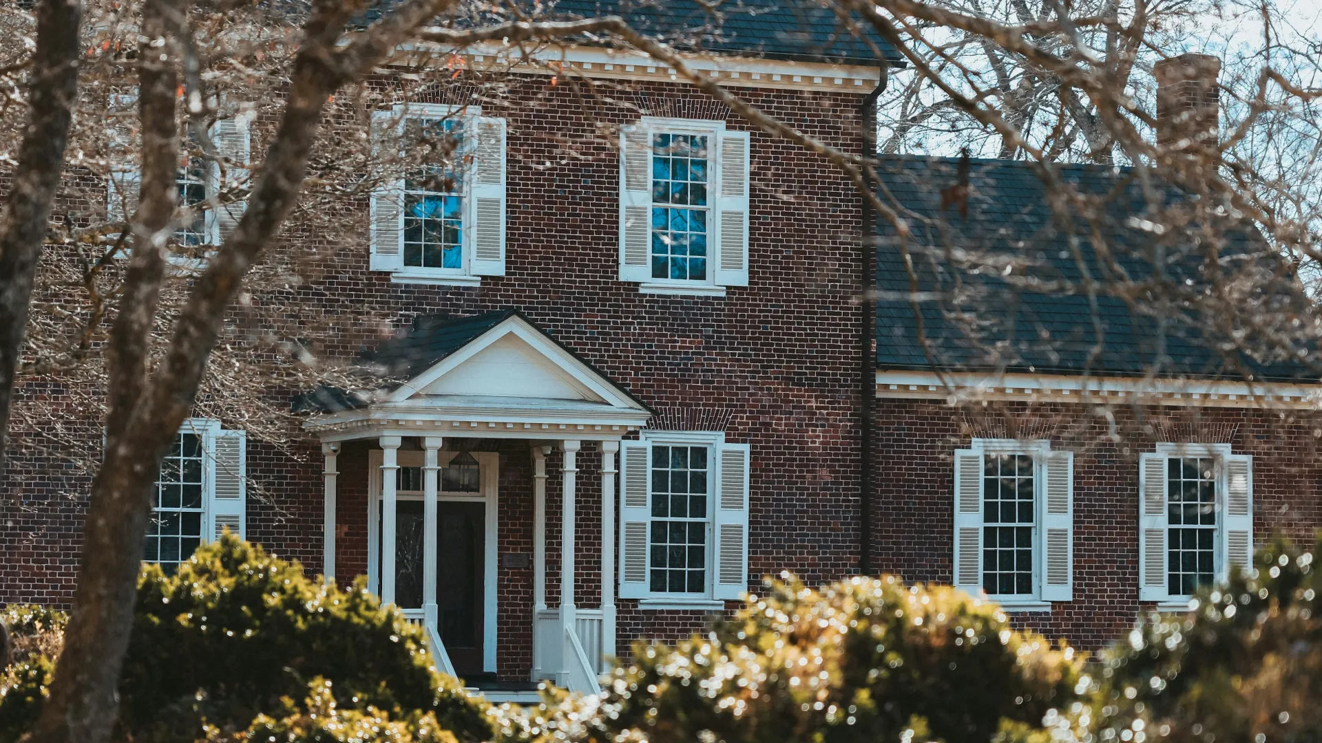 Historic brick house with white shutters and front porch surrounded by leafless trees and bushes in winter.