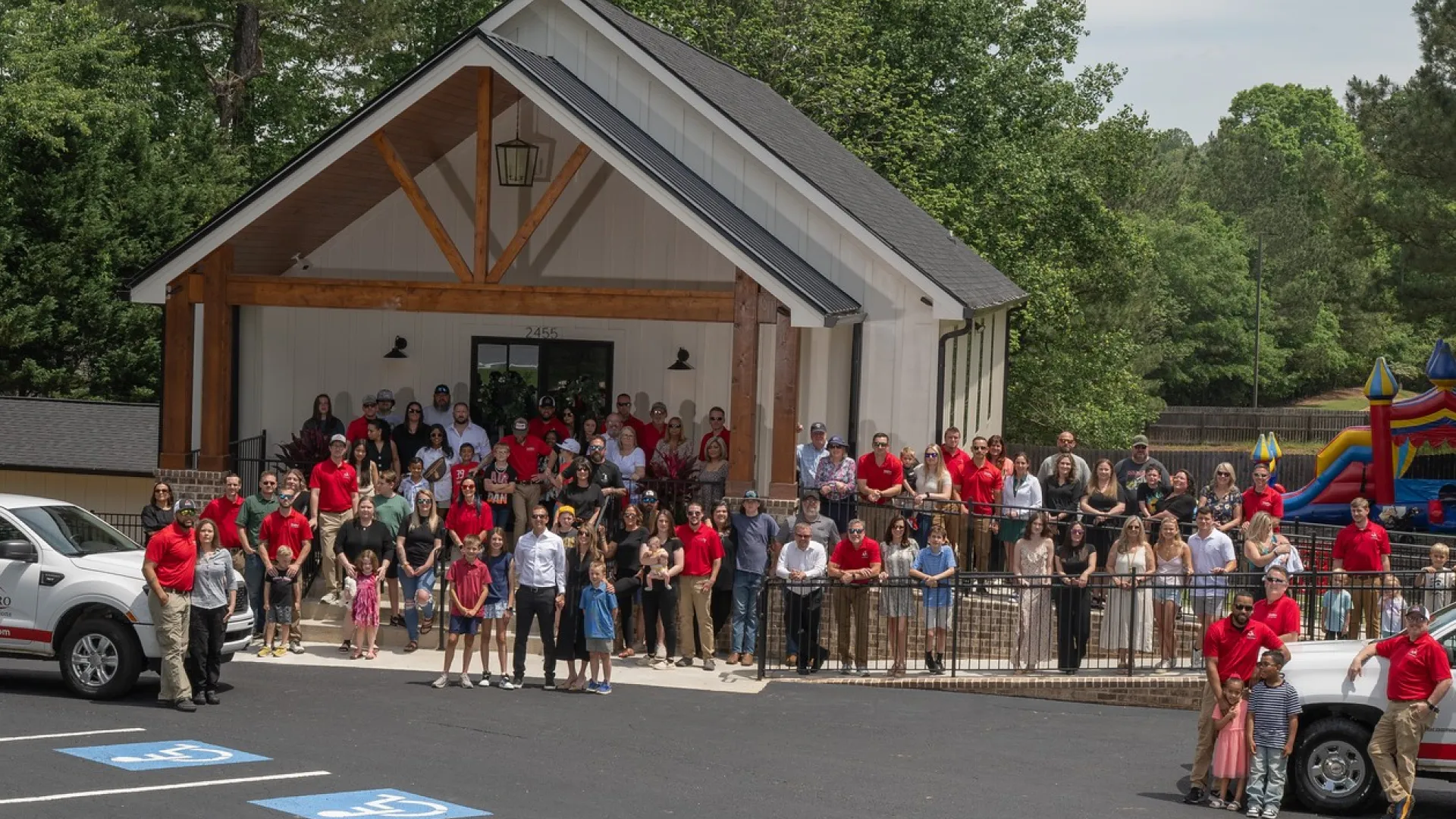 Large group of people gathered outside a modern building with two white trucks in a parking lot surrounded by trees and a bounce house.