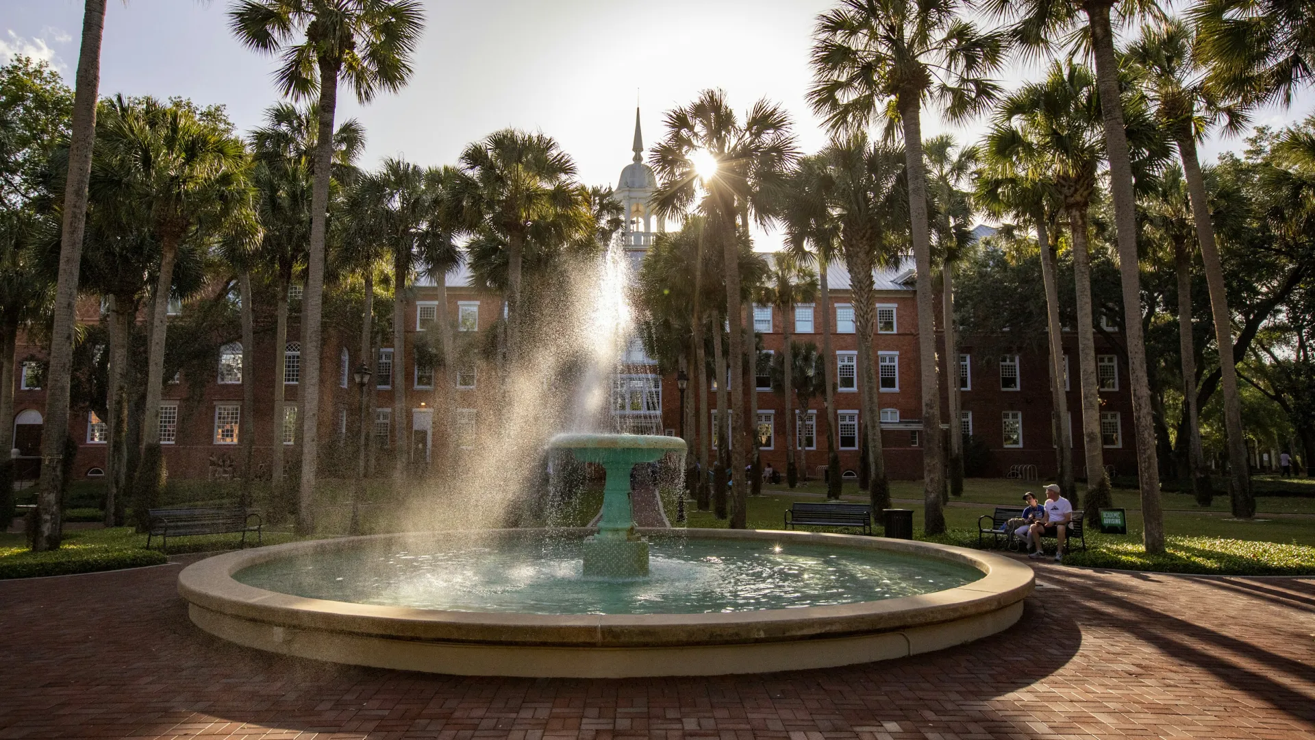Sunlit fountain spraying water in a courtyard with palm trees and a historic brick building in the background.