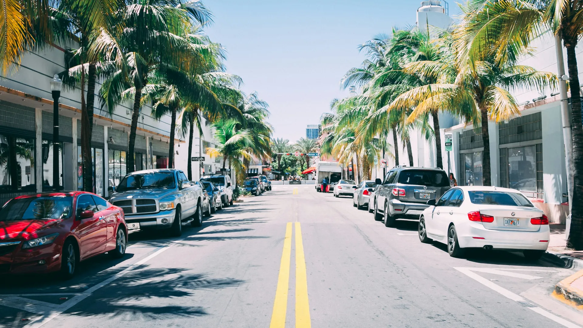 Sunny street lined with palm trees and cars parked along sidewalks in an urban tropical setting.