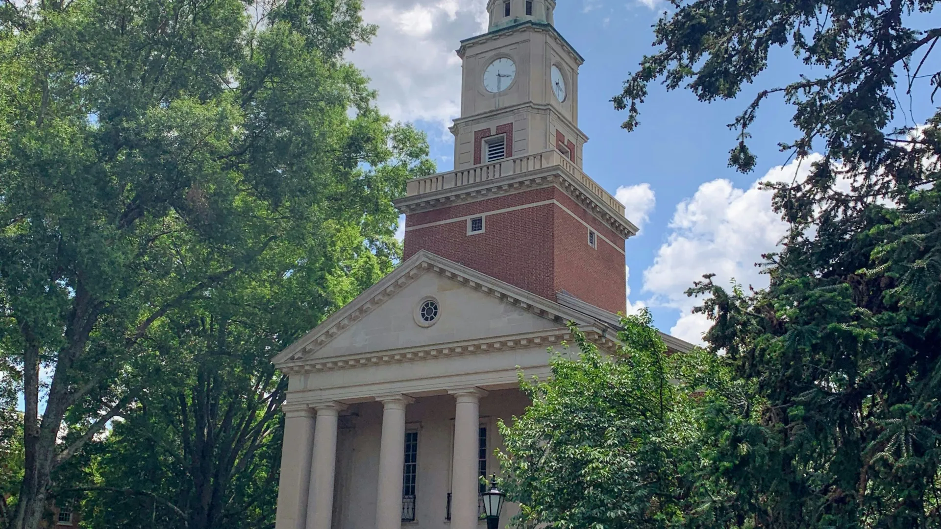 Historic church with tall steeple, clock tower, white columns, surrounded by lush greenery on a sunny day