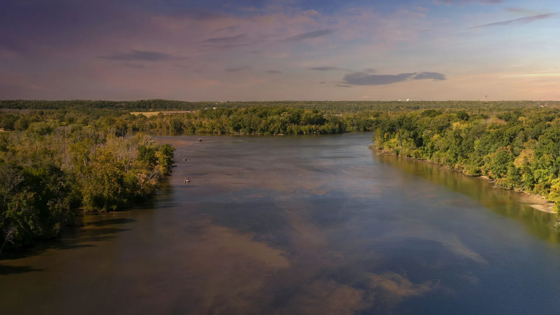 Wide river flowing through dense green forest under a partly cloudy sky at sunset or sunrise.