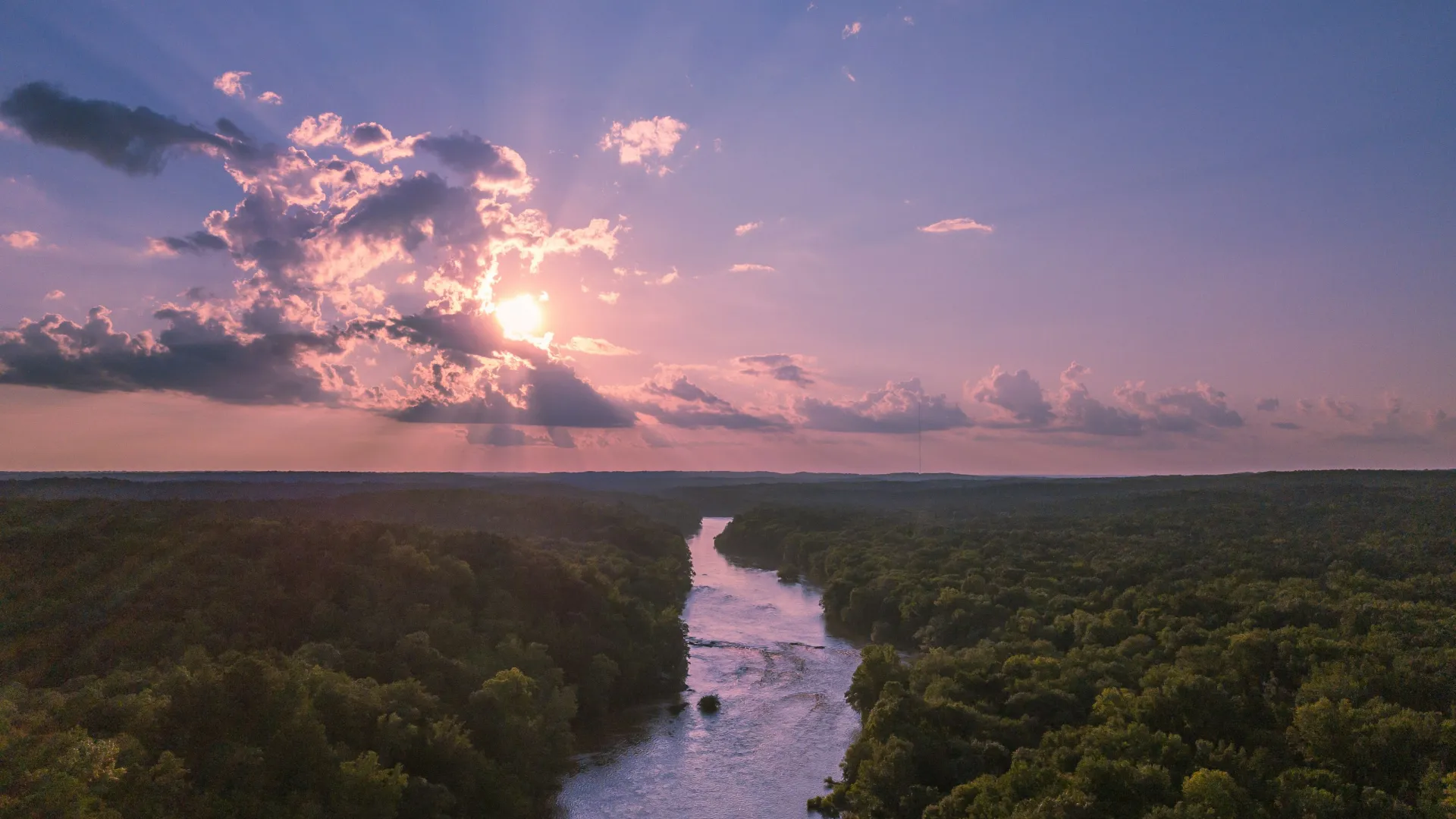 River flowing through dense forest under a purple sunset sky with scattered clouds and sun rays.