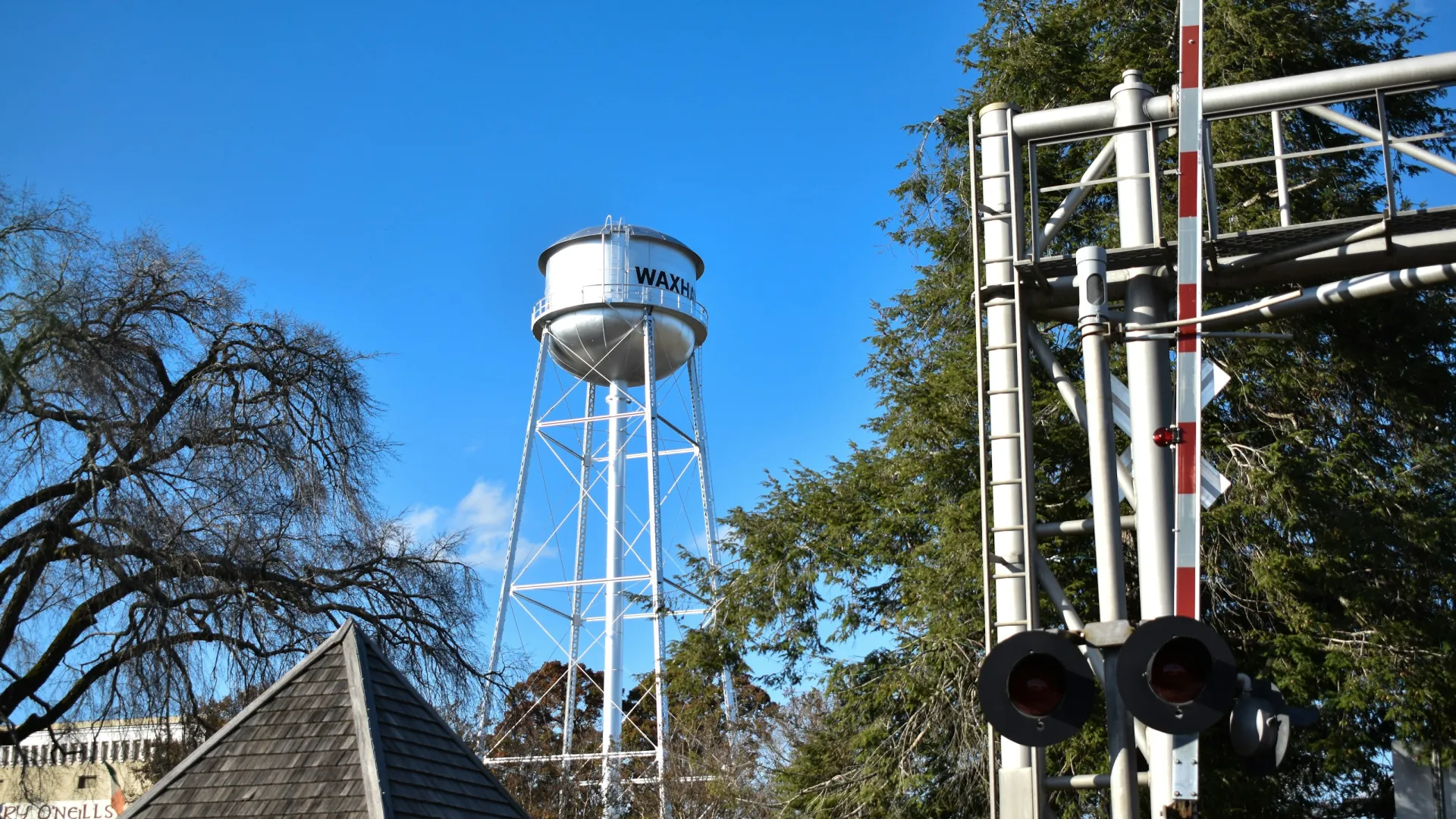 Tall white water tower labeled Waxhaw against a clear blue sky with trees and railroad crossing signals nearby