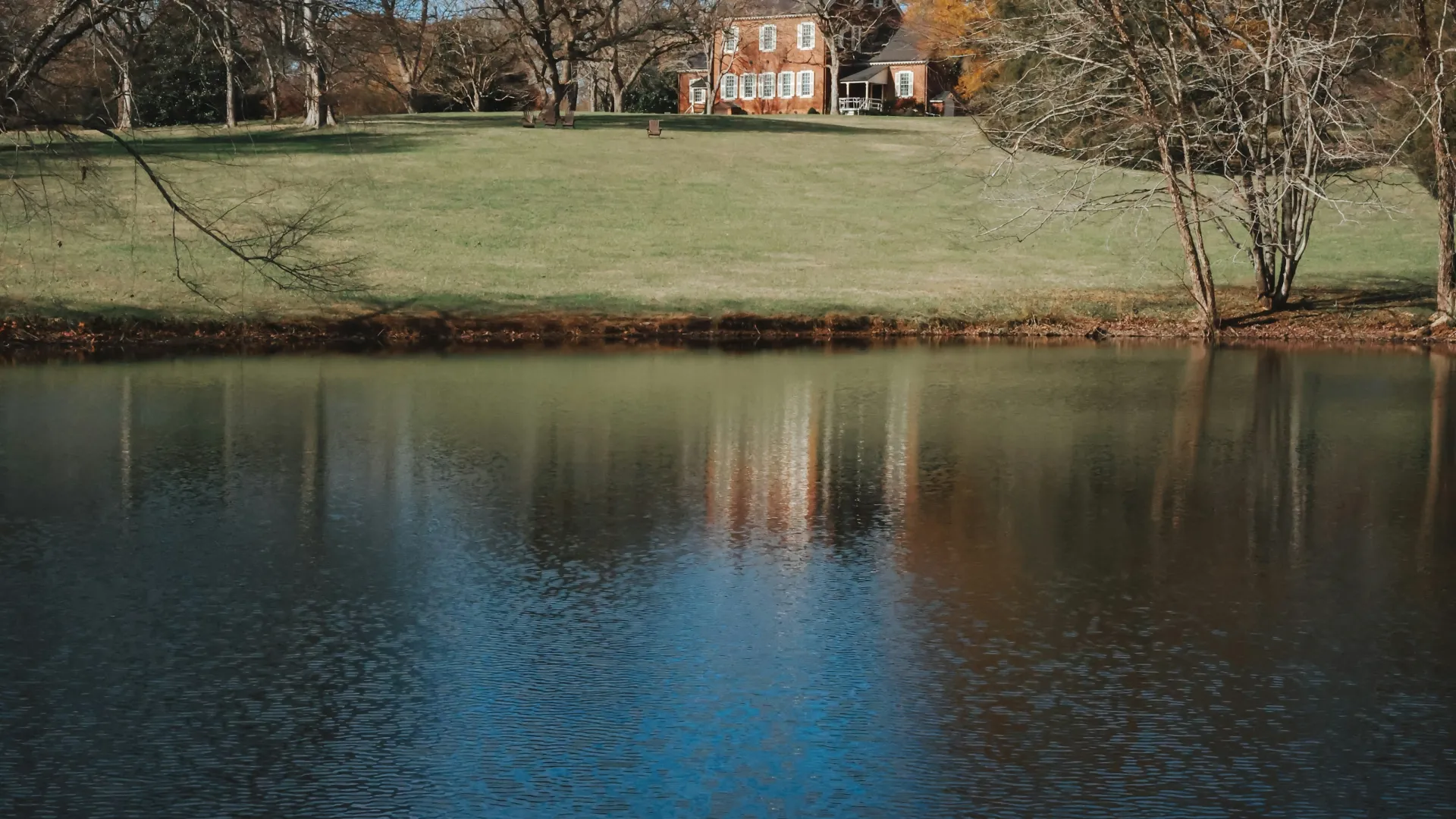 Historic brick house on a grassy hill beside a calm lake with autumn trees and blue sky reflected in the water.