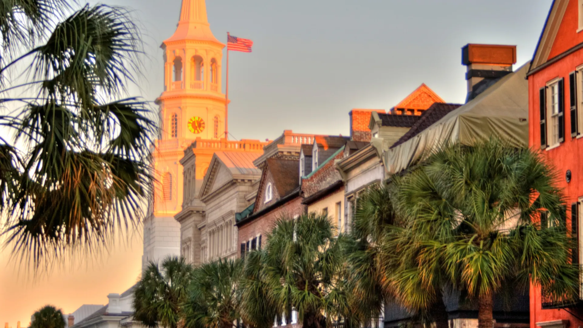 Historic Charleston street with palm trees, colorful buildings, and a white church steeple at sunset.