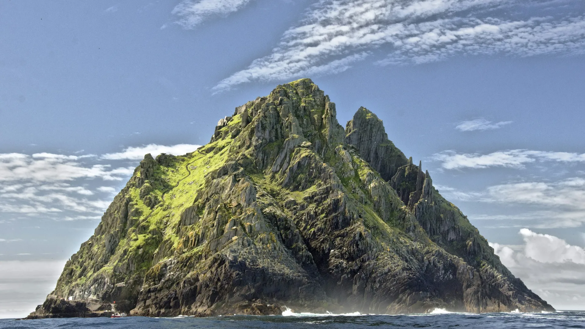 Rocky island with steep cliffs covered in green vegetation surrounded by ocean under partly cloudy sky