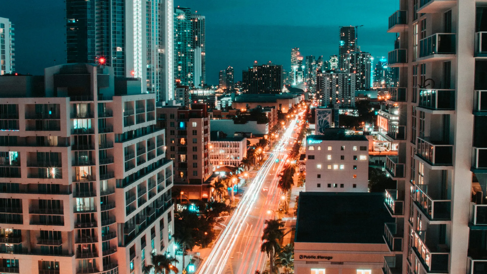 Night cityscape with light trails on a busy street flanked by modern high-rise buildings and palm trees.