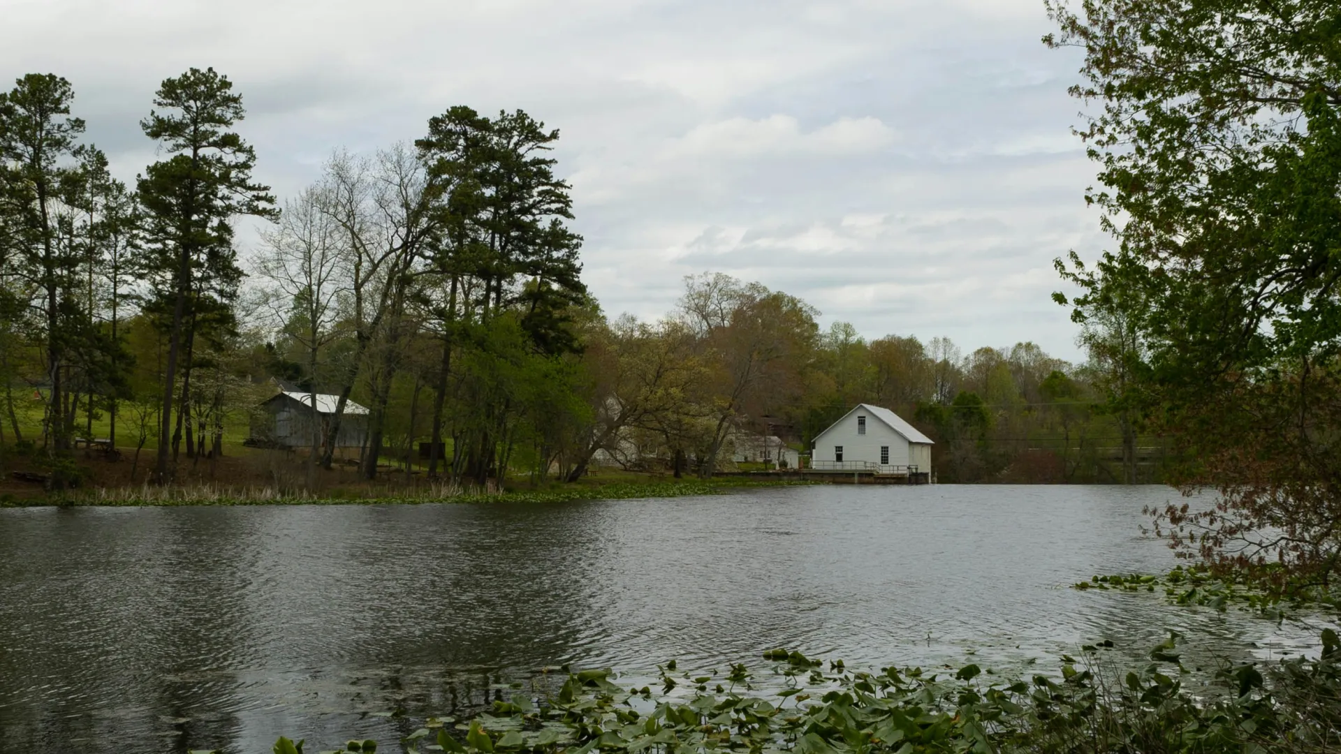 Calm lake surrounded by trees with a white house and other buildings near the water under a cloudy sky