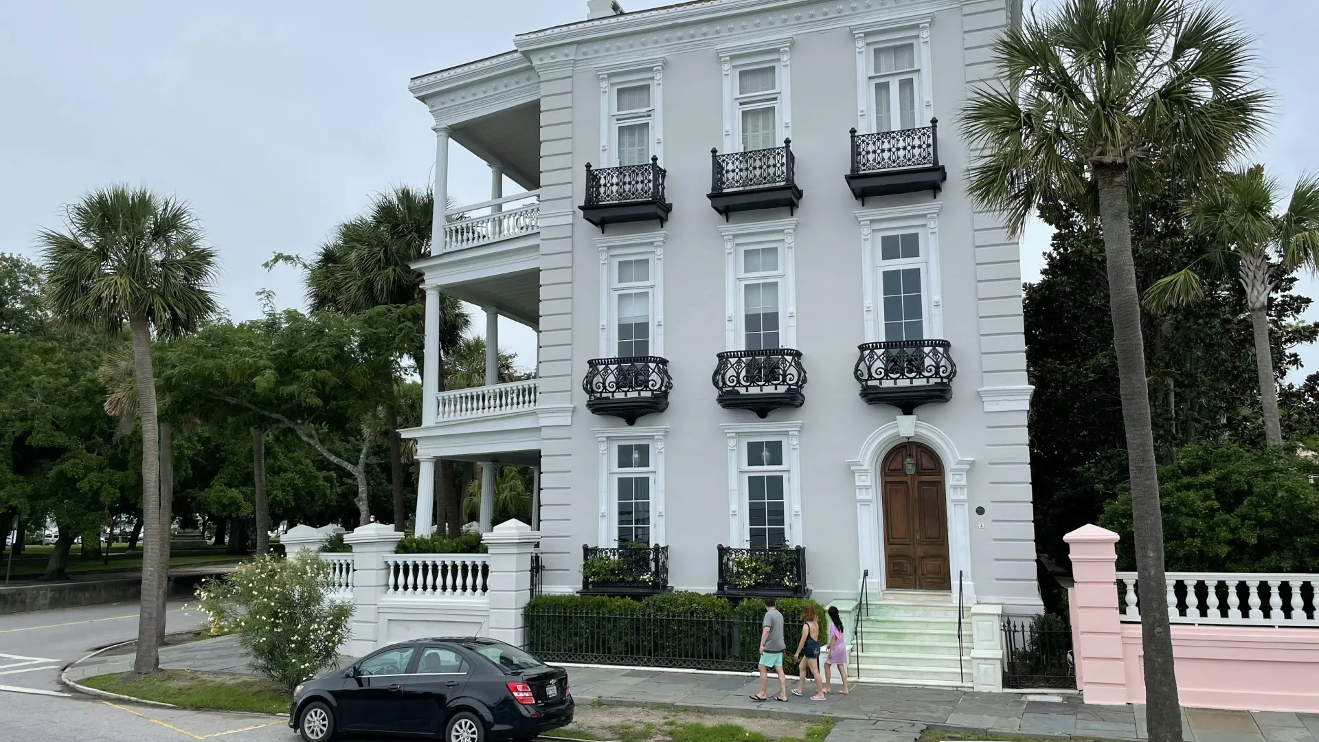 Elegant white three-story historic house with black wrought iron balconies and palm trees on a cloudy day.
