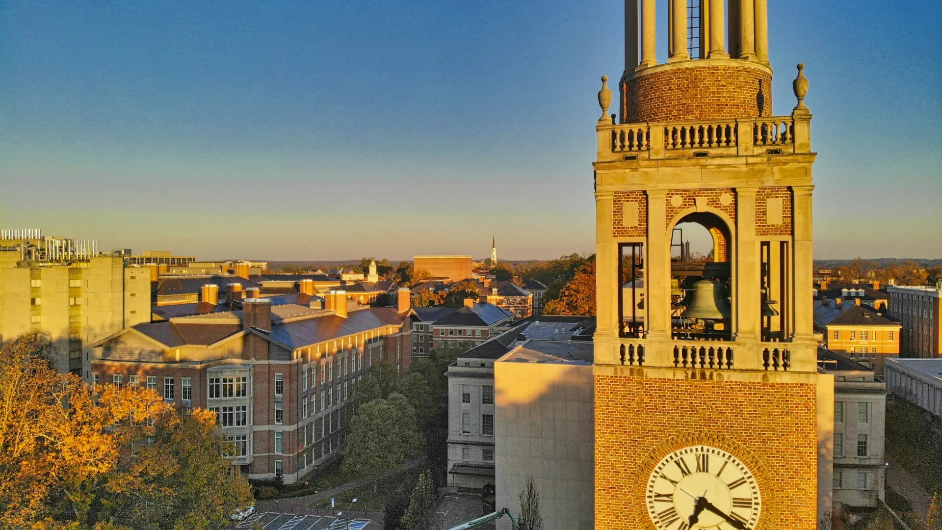 Bell tower with a large clock overlooking a university campus bathed in golden sunlight during sunset.