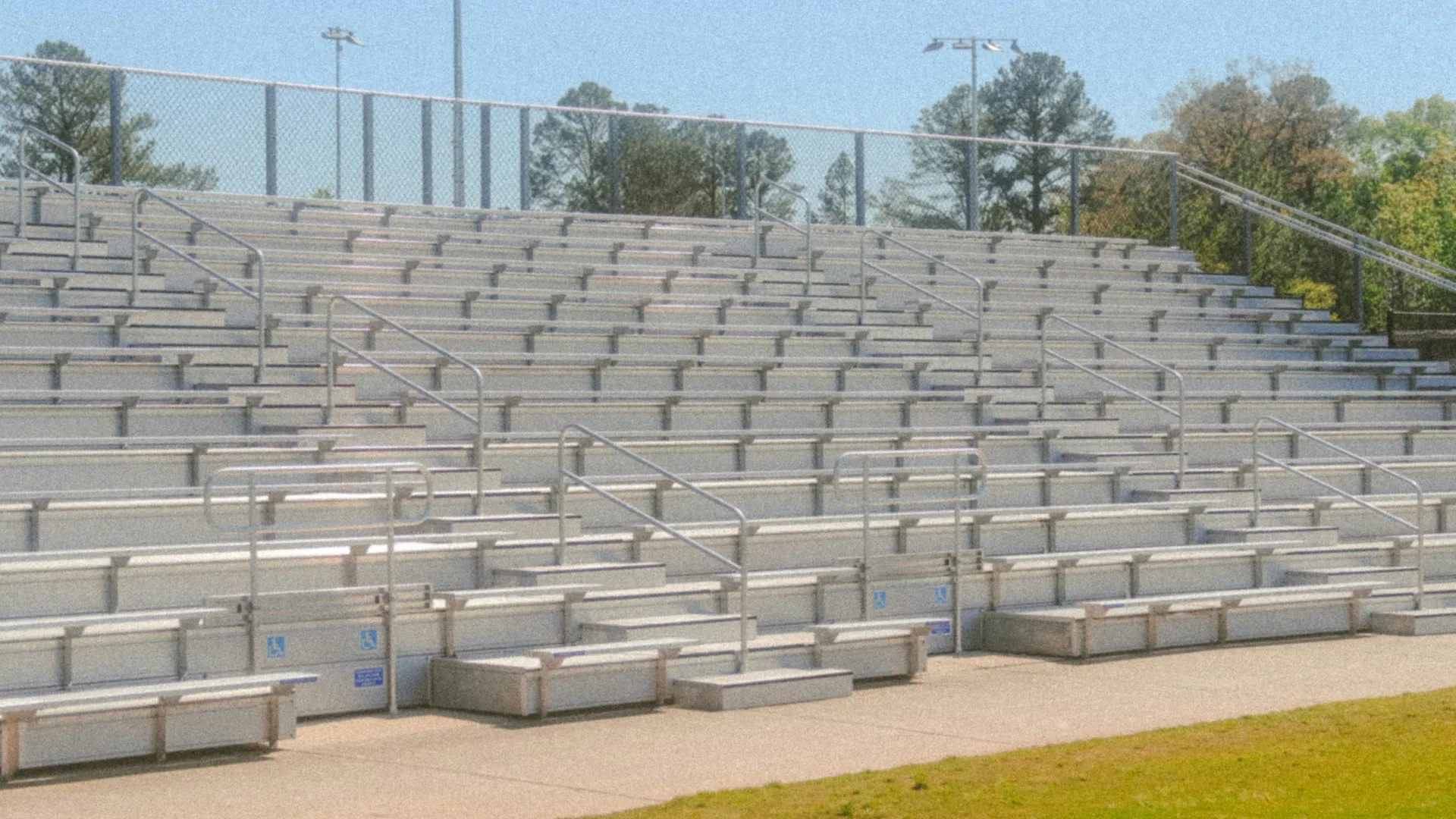 Empty outdoor stadium bleachers with metal railings overlooking a green grass field under clear sky