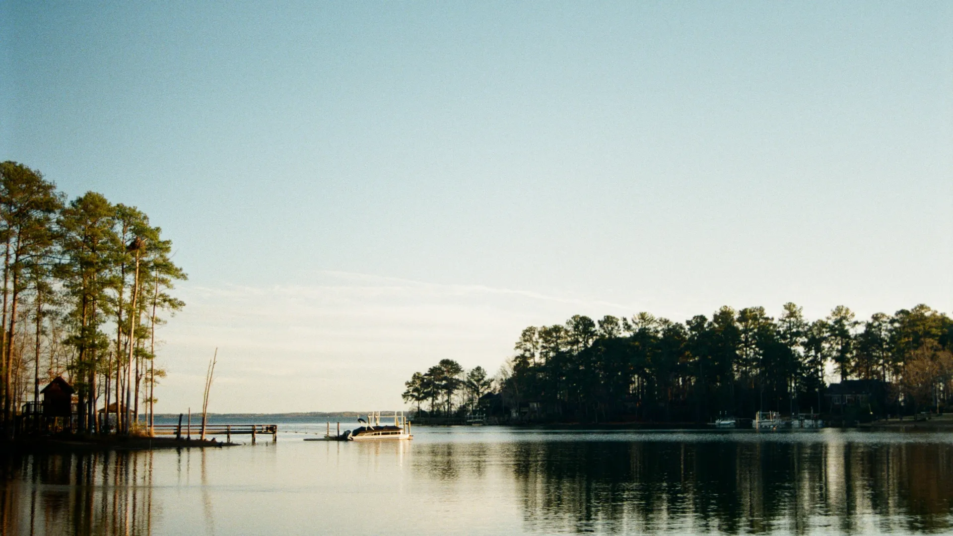 Calm lake with boats docked near tree-lined shore under clear blue sky at sunset.