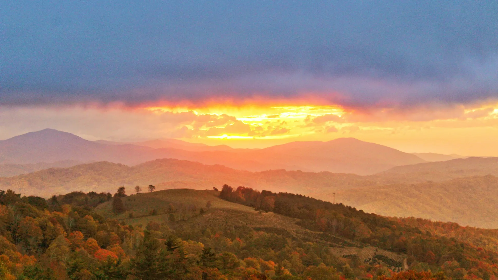 Sunset over rolling hills and autumn forests with layered mountains under a cloudy blue sky.
